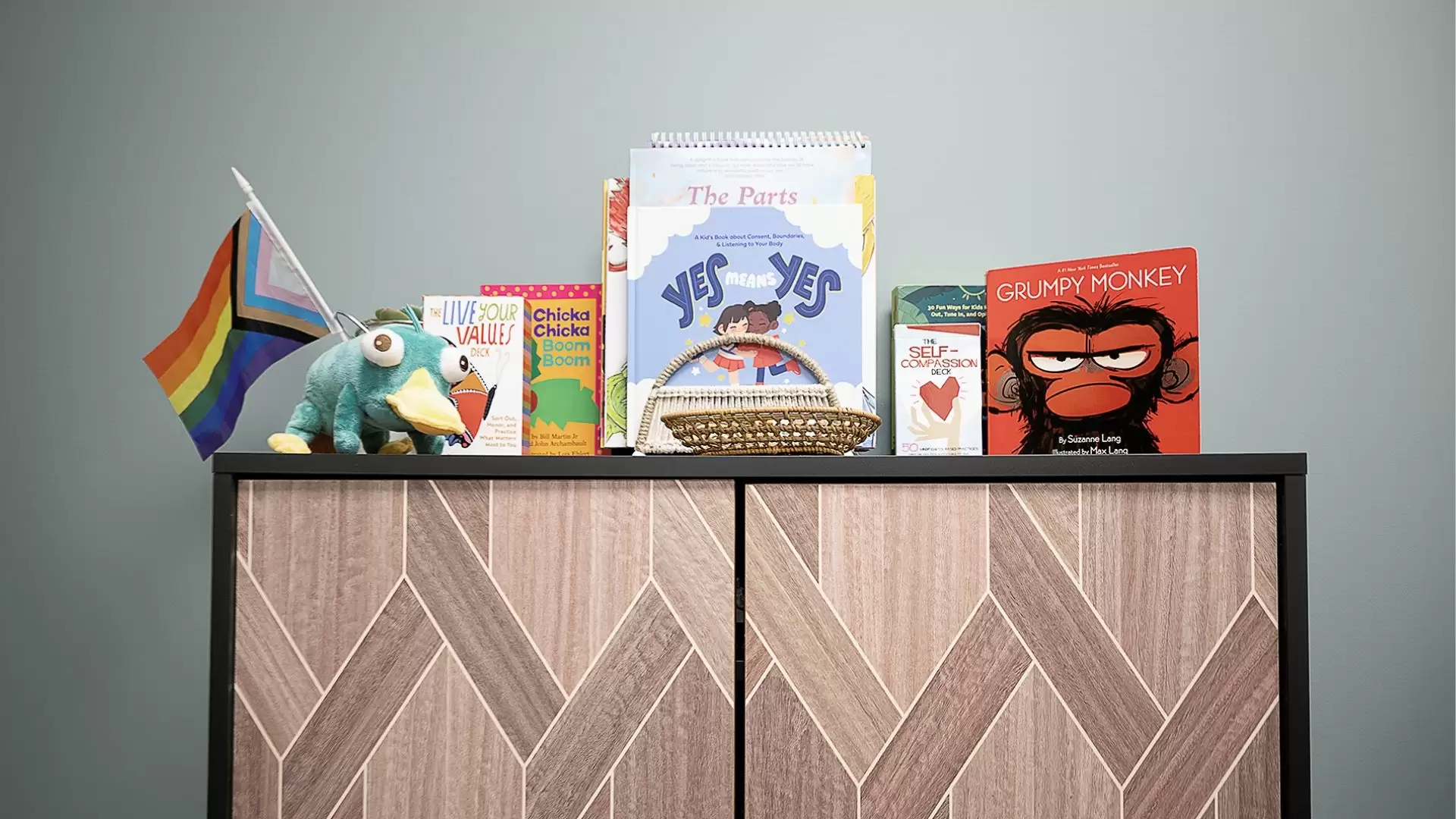 Books for children and youth, a platypus stuffed animal and a progress pride flag on top of a brown wooden cabinet with black border.