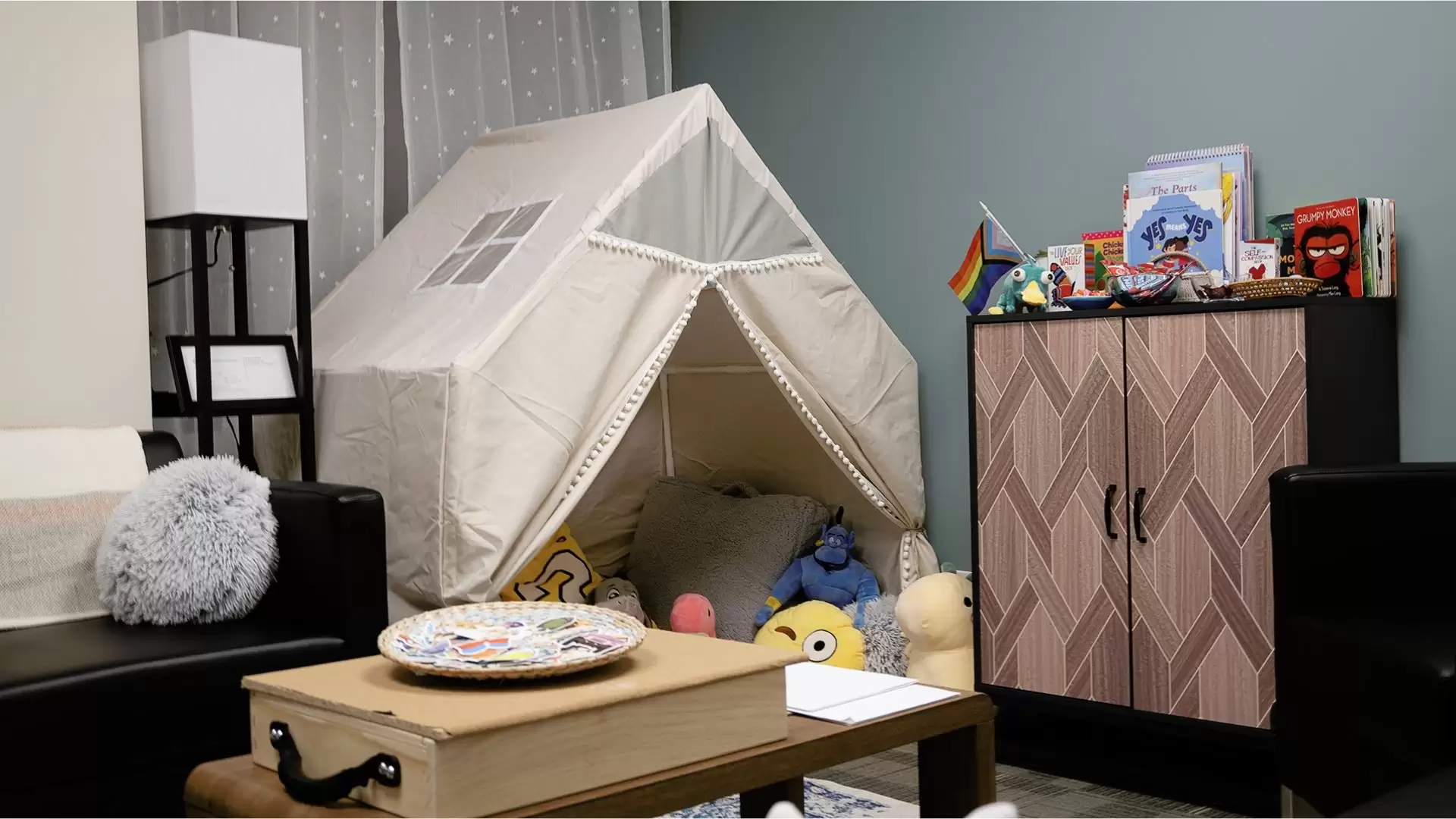 Youth counsellor office with a small cabin style tent filled with plus toys and pillows. A cabinet with children and youth books displayed on the top sits against the wall with a small table and chair in the middle of the office.
