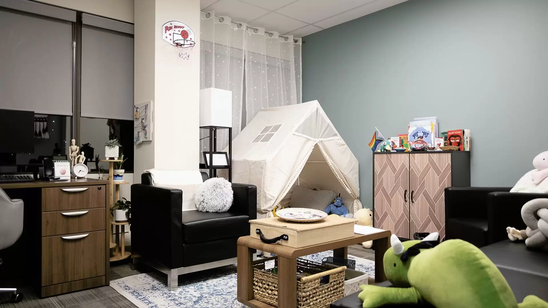 Youth counsellor office with a small cabin style tent filled with plus toys and pillows. A cabinet with children and youth books displayed on the top sits against the wall with a small table and chair in the middle of the office. A desk and computer are against the wall with windows with the blinds pulled halfway down.