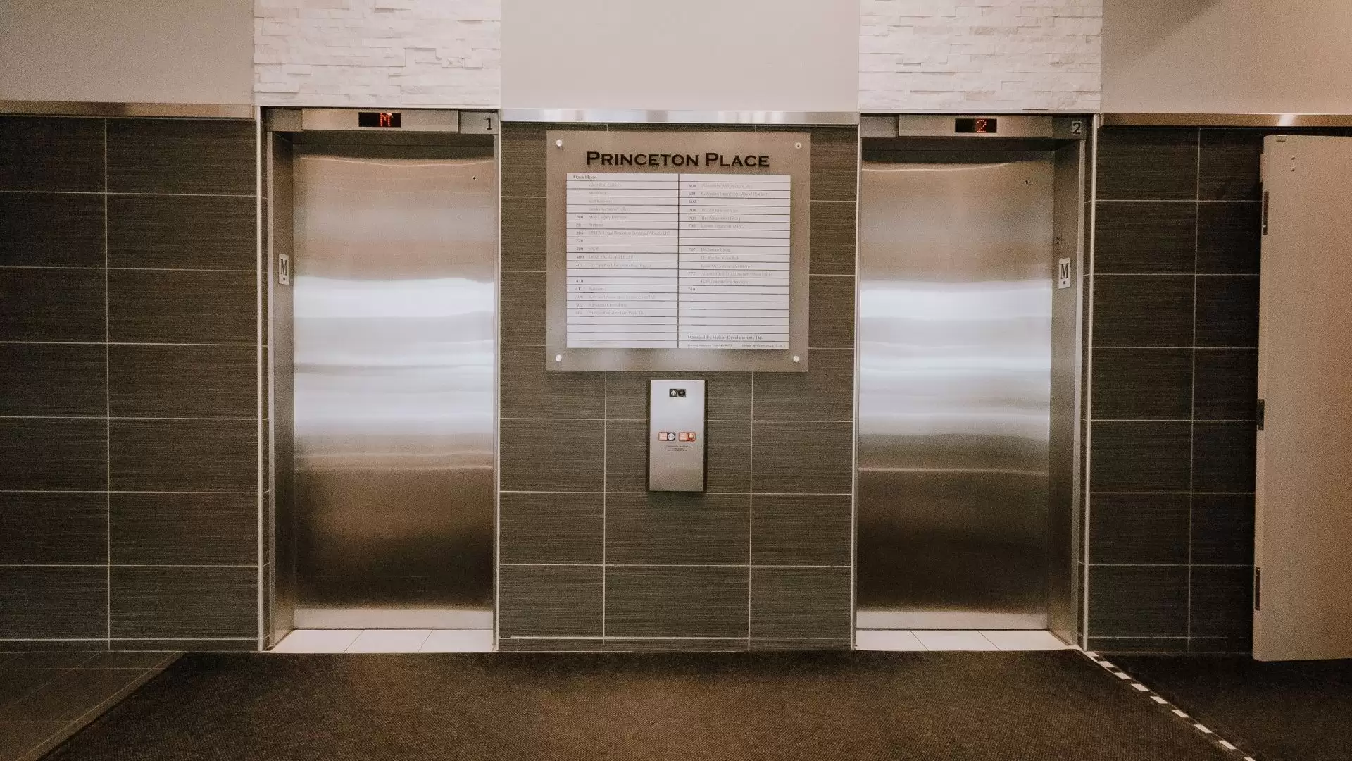 Lobby elevators with grey and neutral walls. The directory sign in the centre has text that says "Princeton Place" with elevator call buttons below.