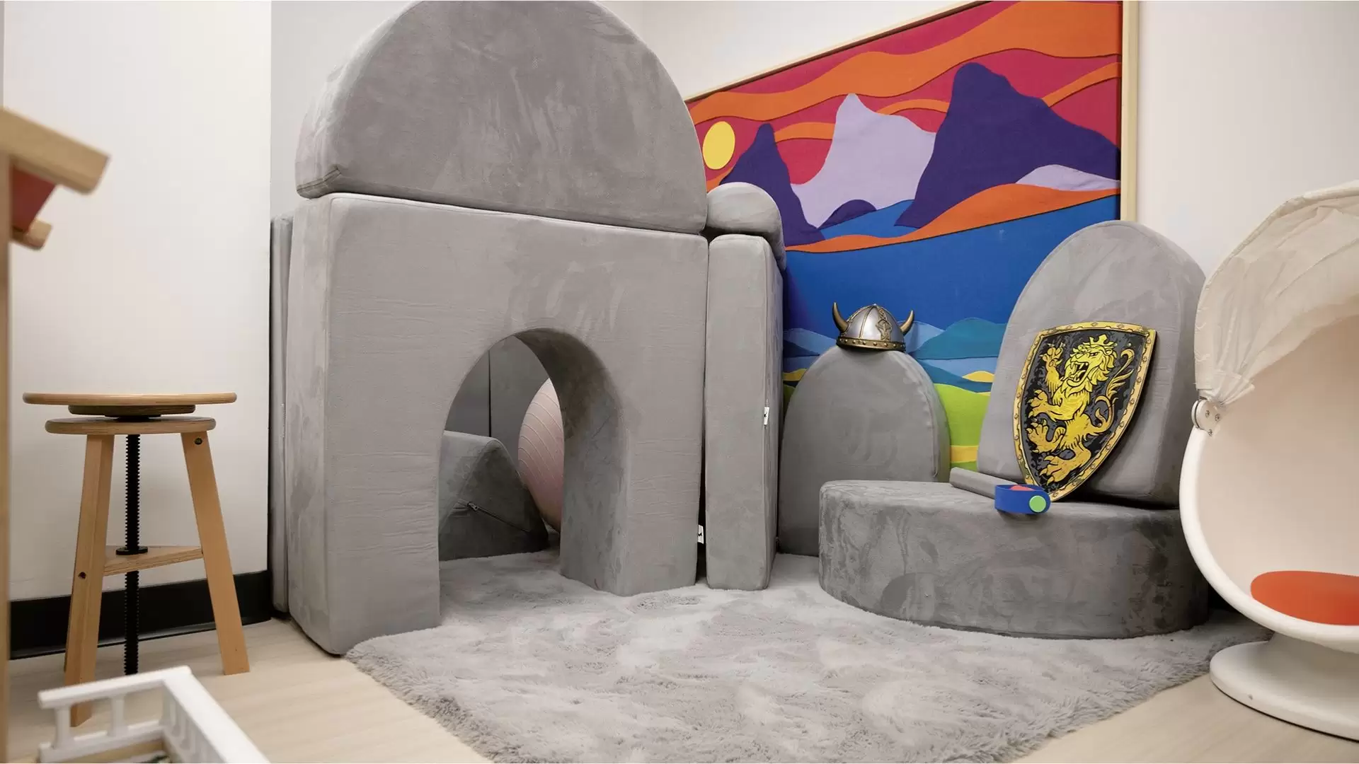 Playroom with large grey pillows in a castle play area with toy shield, sword, and hat on a grey carpet. A colourful felt art board is against the white wall and stools and chairs off to the side.