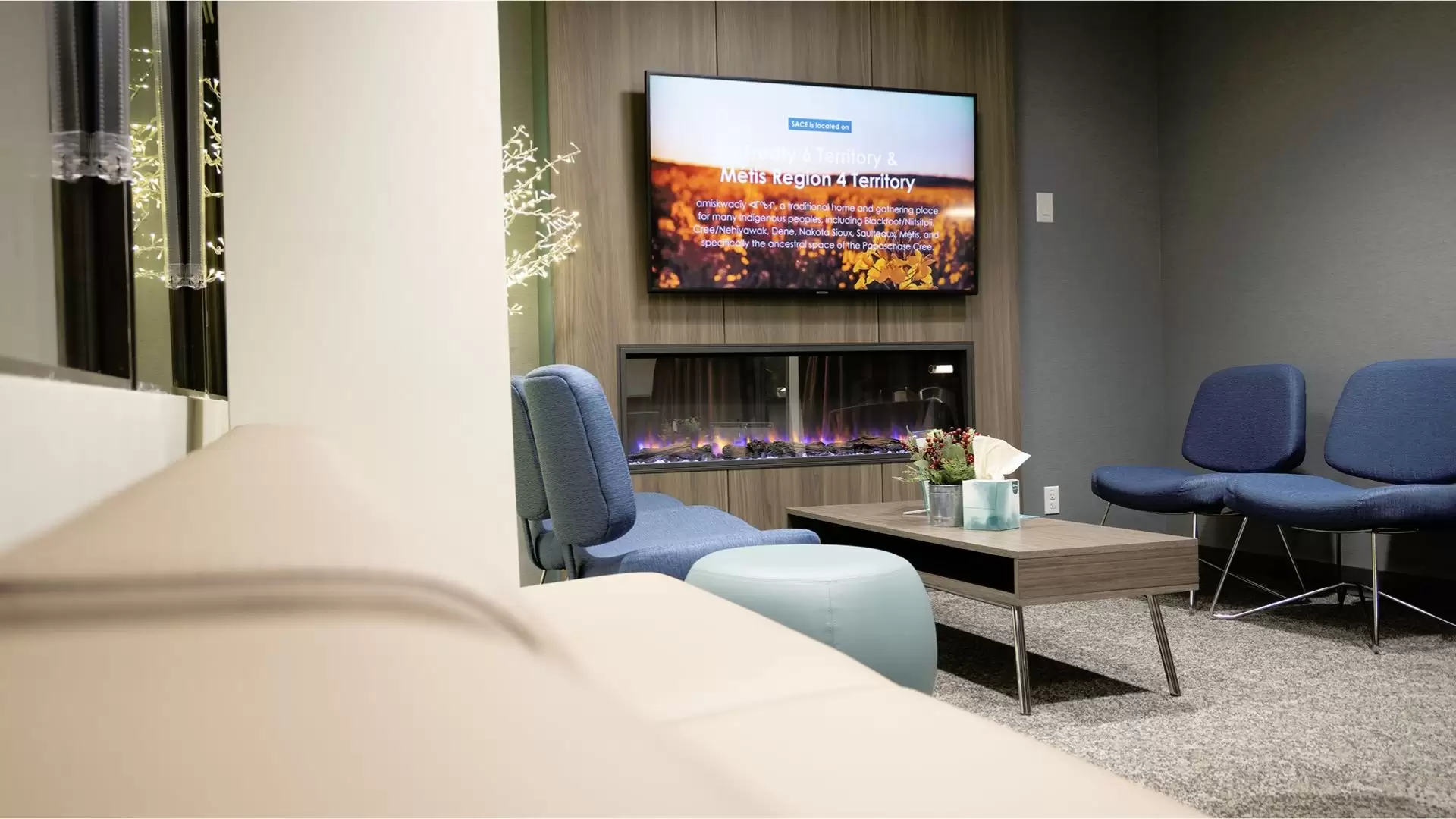 SACE reception waiting area at night with dark chairs, stool, table, and electric fireplace with flat screen TV.
