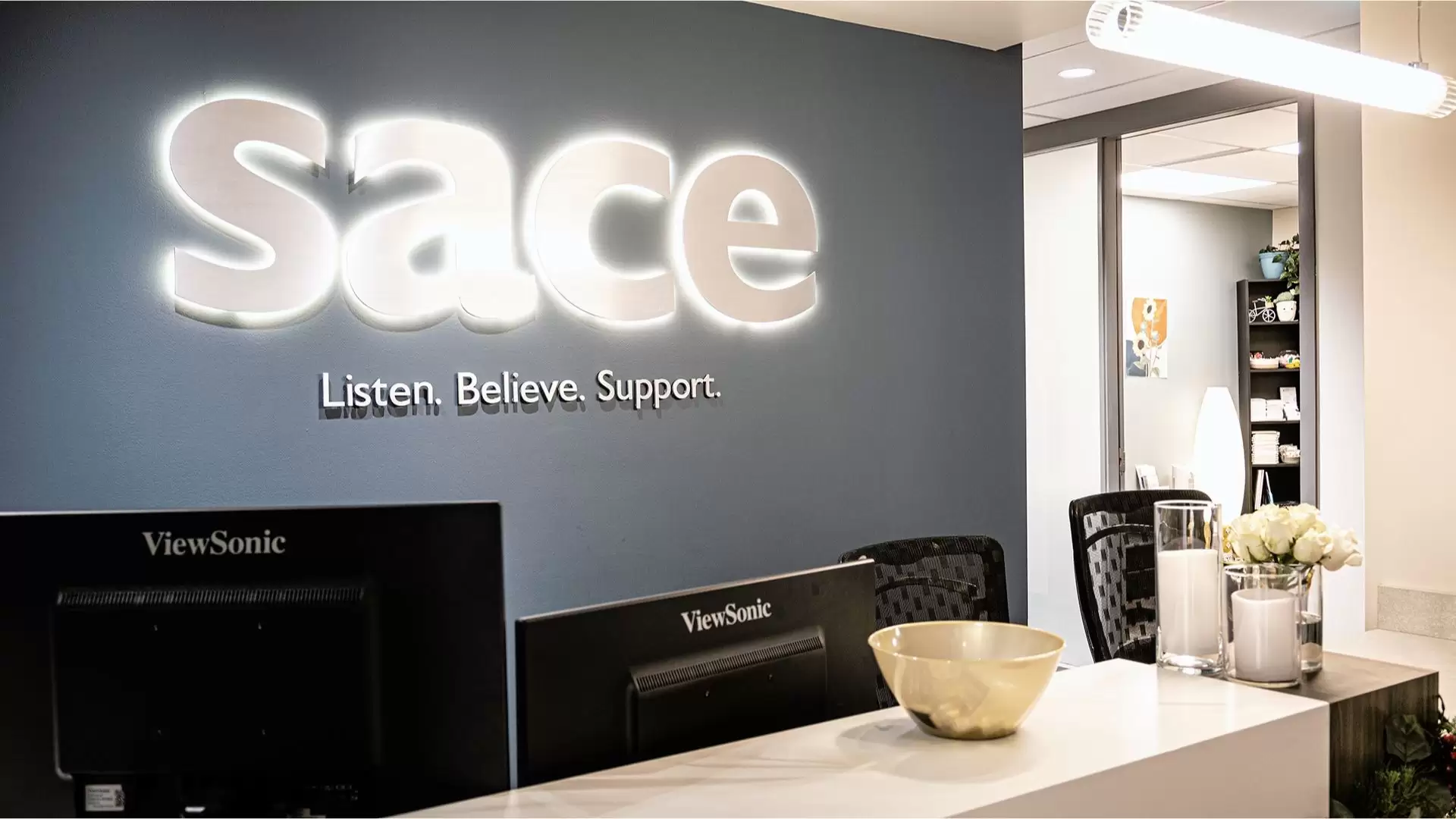 SACE office reception desk with a dark blue painted wall and backlit SACE sign with "Listen. Believe. Support." sign below. Two black monitors and two black office chairs are behind the desk. Candles and white flowers sit on the middle of the desk in glass containers. An office can be seen beyond the front desk.
