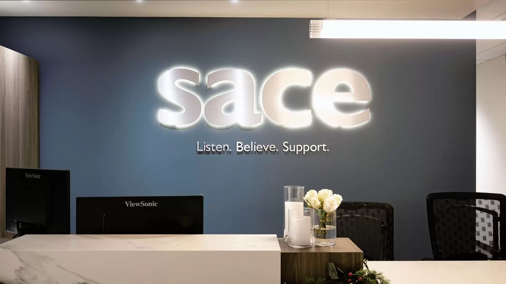 SACE office reception desk with a dark blue painted wall and backlit SACE sign with "Listen. Believe. Support." sign below. Two black monitors and two black office chairs are behind the desk. Candles and white flowers sit on the middle of the desk in glass containers.