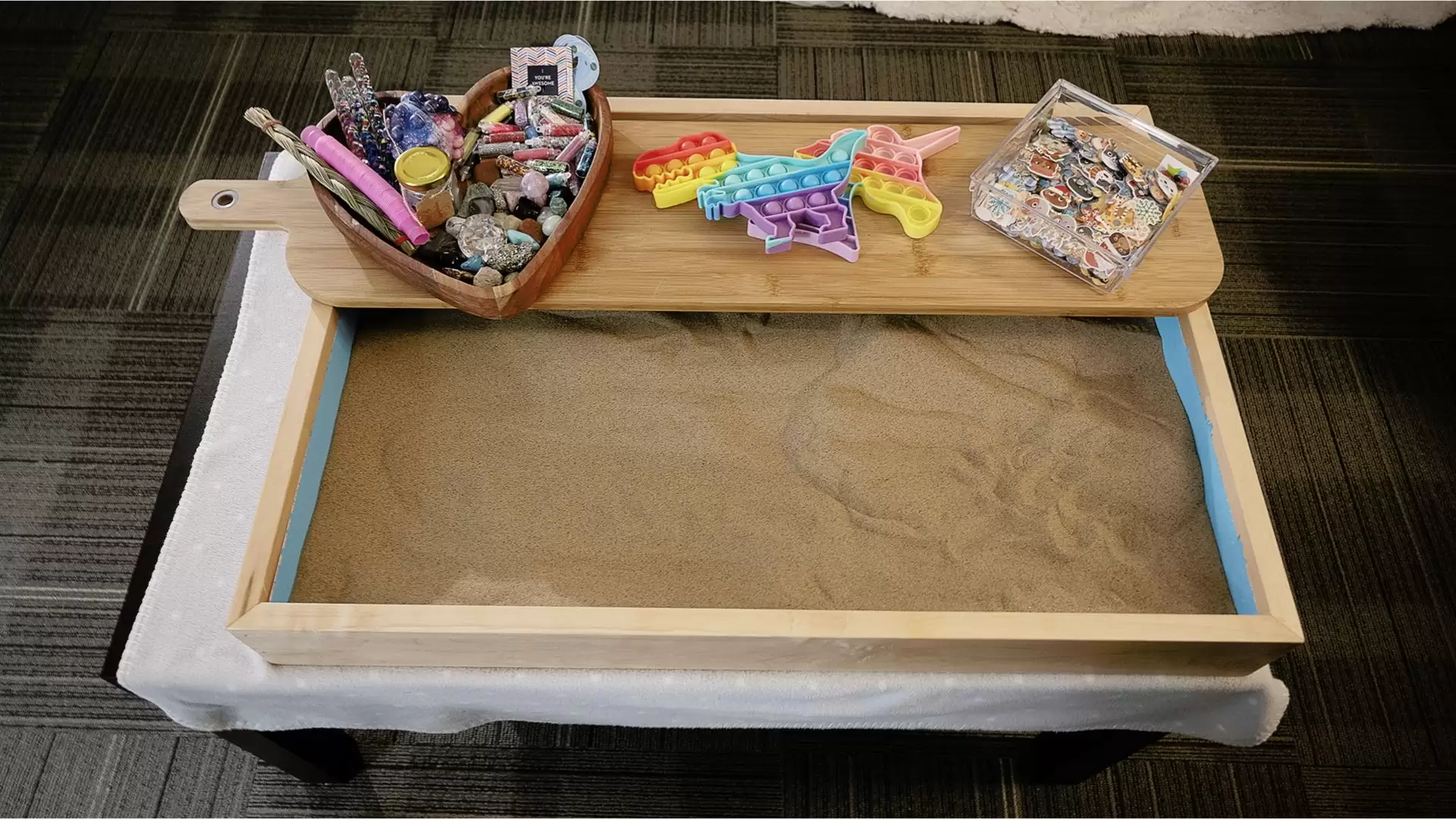 Sandbox tray filled with sand. A narrow wood cutting board is placed across the top with a heart shaped wooden box filled with stones, rocks and decorative objects, rainbow coloured dinosaur and unicorn push pop bubble fidget sensory toys, and a plastic tray of animal shaped objects.