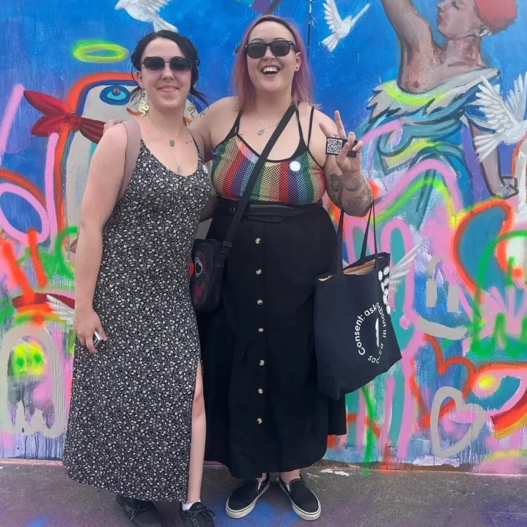 Two SACE educators stand smiling in front of a colourful graffiti wall at Edmonton Pride 2024